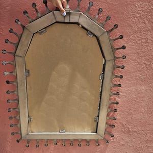 May include: A decorative mirror with a distinctive sunburst design. The mirror has a rectangular shape with a metal frame and droplet-shaped accents around the edge. The back of the mirror is visible, showing a brown backing.