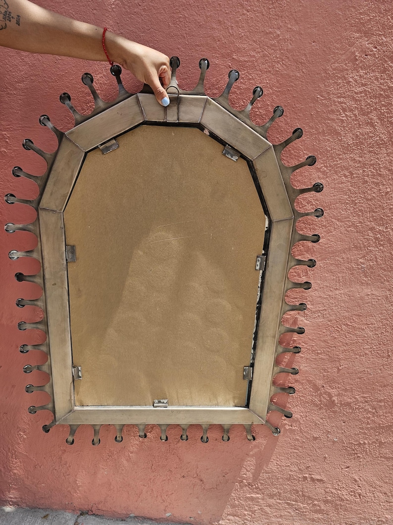 May include: A decorative mirror with a distinctive sunburst design. The mirror has a rectangular shape with a metal frame and droplet-shaped accents around the edge. The back of the mirror is visible, showing a brown backing.