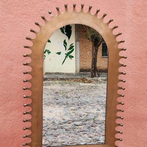 Puede incluir: Un espejo de pared arqueado con un marco de bronce metálico y un borde festoneado decorativo. El marco está adornado con una serie de pequeñas protuberancias esféricas espaciadas uniformemente a lo largo de todo el perímetro exterior, creando una silueta de corona.