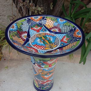 May include: A vibrant, hand-painted ceramic birdbath with a pedestal base. The birdbath features a colorful floral design in shades of blue, orange, green, and red. The pedestal is cylindrical and also decorated with matching patterns.