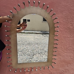 May include: A decorative mirror with a distinctive frame. The frame is a light metallic colour with a scalloped edge, each scallop topped with a small, round, silver-coloured ball. The mirror has an arched top and reflects an outdoor scene.