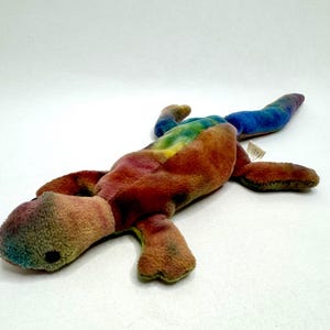 May include: A tie-dye plush toy in the shape of a lizard. The toy is a mix of orange, yellow, green, blue, and purple.