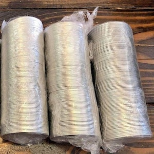 May include: Three rolls of silver-colored aluminum foil, each wrapped in clear plastic, are stacked on a wooden surface.