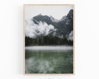 May include: A framed landscape print featuring a misty mountain scene. The image shows a lake in the foreground, with a forest and mountains in the background. The mountains are partially obscured by clouds. The frame is light-colored wood.