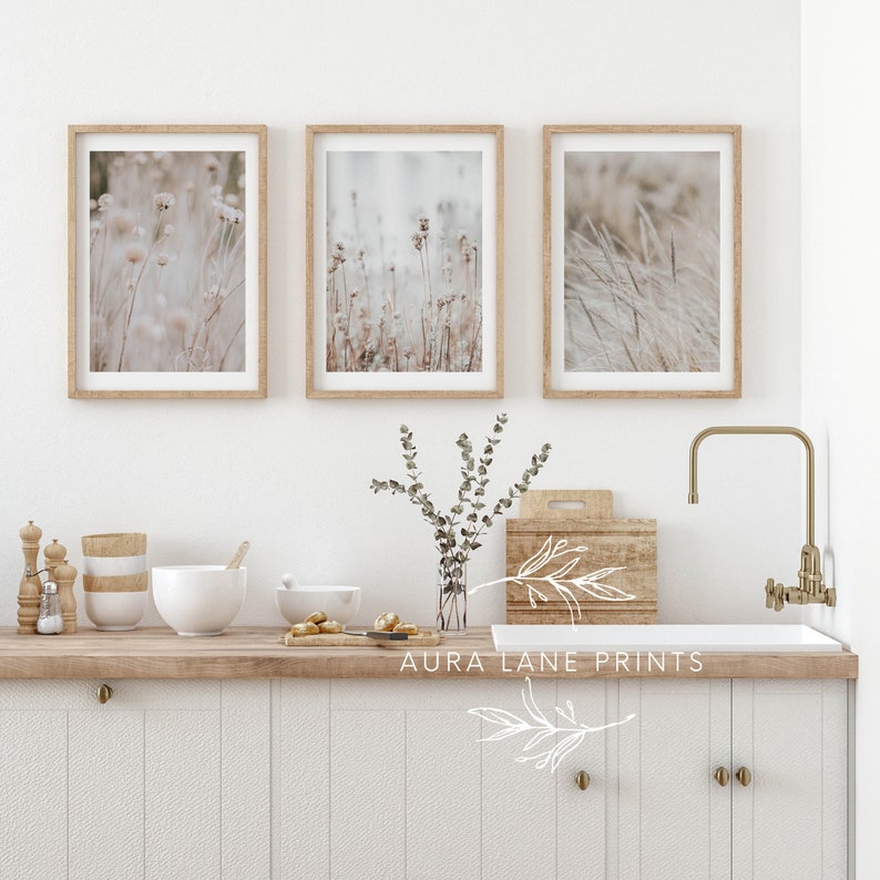 May include: Three framed prints of muted beige and brown grasses and flowers in a minimalist kitchen setting. The prints are hung on a white wall above a wooden countertop with a vase of eucalyptus branches and a wooden cutting board.
