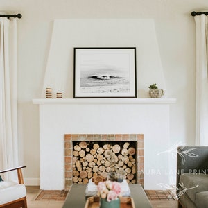 May include: A black and white photograph of a wave crashing on a beach hangs above a white fireplace with a brick surround. The fireplace is filled with stacked logs. A gray sofa with a white throw pillow is in the foreground.