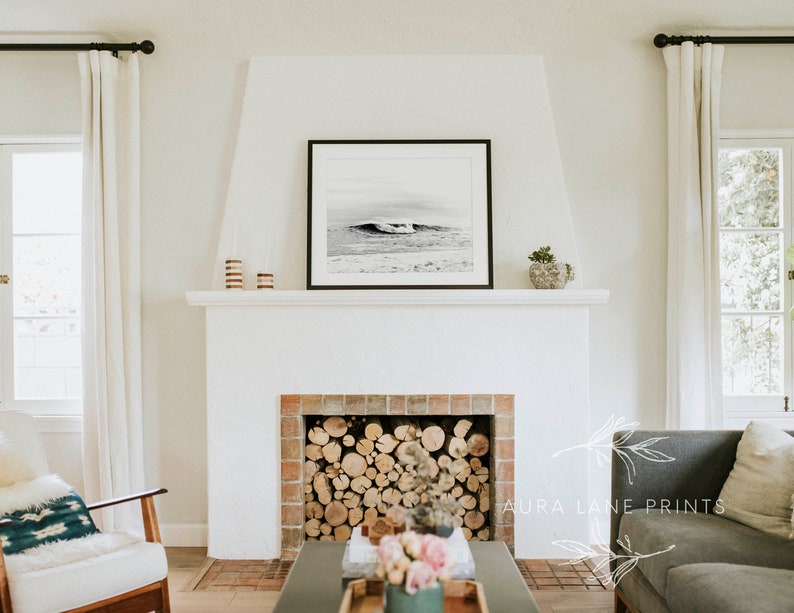 May include: A black and white photograph of a wave crashing on a beach hangs above a white fireplace with a brick surround. The fireplace is filled with stacked logs. A gray sofa with a white throw pillow is in the foreground.
