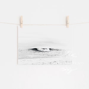 May include: A black and white photograph of a wave crashing in the ocean. The photo is hanging on a white wall with a string and clothespins.