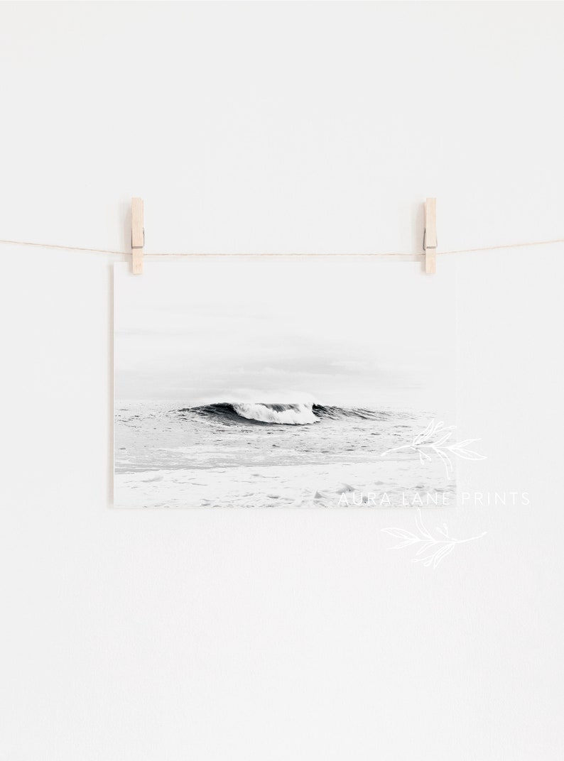 May include: A black and white photograph of a wave crashing in the ocean. The photo is hanging on a white wall with a string and clothespins.
