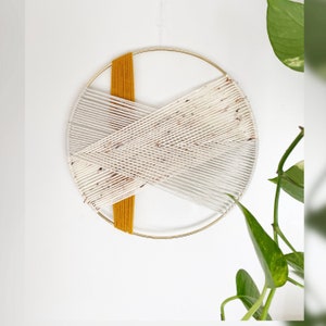 May include: A white and gold macrame wall hanging with a geometric design. The macrame is made with white and yellow yarn and is woven around a gold metal hoop.