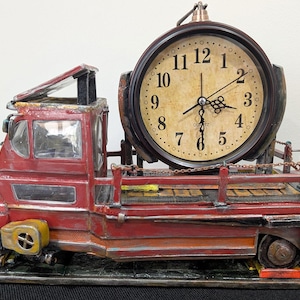 May include: A red truck-shaped table clock with a round, cream-colored face featuring black Arabic numerals and ornate hands. The truck has a flatbed design with a small chain railing, a weathered finish, and the text CLAY WEYLAND in the corner.