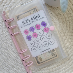 May include: A clear plastic binder with a pink zipper and a pink ring binder mechanism. The binder contains a printed sheet with a savings challenge game. The game has a grid of hexagons with dollar amounts written in each hexagon. Some of the hexagons are filled in with purple and pink. The sheet also has the text "$25 Mini Savings Challenge" and "NessaBudgets".