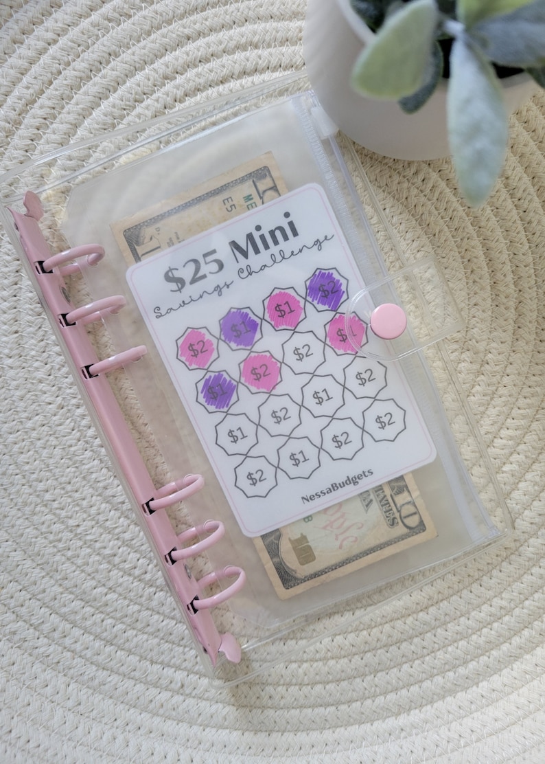 May include: A clear plastic binder with a pink zipper and a pink ring binder mechanism. The binder contains a printed sheet with a savings challenge game. The game has a grid of hexagons with dollar amounts written in each hexagon. Some of the hexagons are filled in with purple and pink. The sheet also has the text "$25 Mini Savings Challenge" and "NessaBudgets".