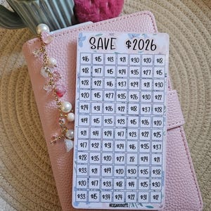 May include: A pink planner with a savings tracker card that reads "SAVE $2026." The card has a grid of boxes with dollar amounts. A charm with pearls and beads hangs from the planner. A decorative strawberry is visible.