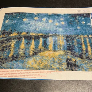 May include: A close-up of a diamond painting kit featuring a starry night scene. The kit includes a canvas with a printed design, a tray of diamonds, and a tool for placing the diamonds. The instructions for the kit are visible at the bottom of the image.