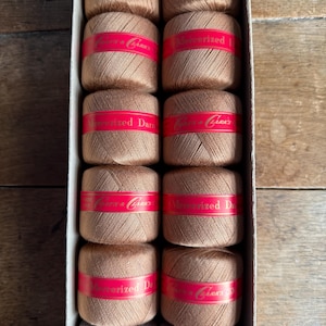 Puede incluir: Una caja de carretes de hilo en varios tonos de marrón y gris. Cada carrete está envuelto con una banda roja con texto blanco. Las etiquetas dicen "Mercerized Darns" y "Clark's".