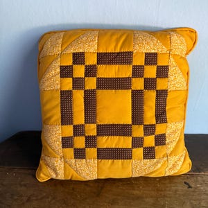 May include: A square quilted throw pillow featuring a patchwork design in mustard yellow and dark brown. The center displays a geometric block pattern with brown fabric featuring small white dots, surrounded by solid yellow and floral yellow fabric.