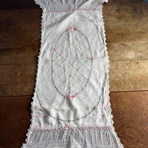 May include: White embroidered table runner with a central oval design. The runner features a diamond pattern, pink and green floral embroidery, and a scalloped edge. The fabric appears to be a lightweight cotton or linen.