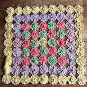 May include: A square textile piece composed of numerous fabric circles in pastel colors: yellow, lavender, pink, and green. The circles are arranged in a grid pattern, creating a textured, floral-like design. The background is a wooden surface.