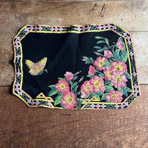 Puede incluir: Un posavasos de fieltro negro con una mariposa amarilla y flores rosas y amarillas impresas en él. El posavasos tiene un borde dorado y verde.
