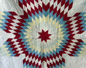 Può includere: Una trapunta con un grande motivo a stella centrale a otto punte composto da piccoli pezzi di tessuto a forma di rombo in rosso, azzurro e giallo pallido. Lo sfondo è bianco con intricate cuciture trapuntate a linee ondulate.