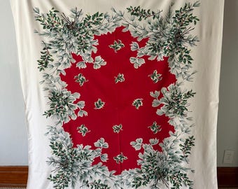 May include: A rectangular tablecloth with a white background and a large central red panel. The red section features scattered green holly leaf motifs. A wide border of pine branches, holly leaves, and pinecones frames the central red area.