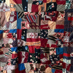 May include: A patchwork quilt constructed from various fabric scraps in shades of deep red, navy blue, black, and tan. The quilt features a mix of patterns including floral prints, geometric shapes, stripes, and abstract designs joined by visible zigzag stitching.