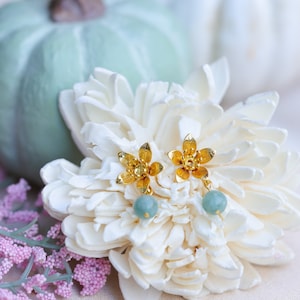 May include: Gold flower earrings with light green gemstone drops. The earrings are displayed on a white flower.
