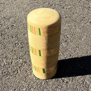 May include: A cylindrical wooden object with a rounded top. The body is light-colored wood with bands of darker wood and green accents. The object is standing upright on a textured gray surface, casting a shadow.