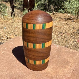 May include: A cylindrical wooden object with a rounded top, featuring alternating horizontal bands of dark brown walnut, light tan wood, and thin strips of vibrant green. The surface is smooth and polished, showcasing the natural grain of the wood.