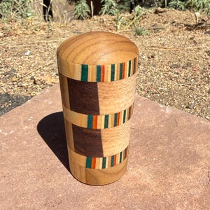 May include: A cylindrical wooden container with a rounded lid, featuring a segmented design of light and dark brown wood panels accented by horizontal bands of thin, multicolored stripes in shades of green, orange, and cream.