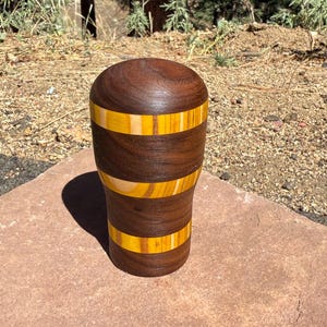 May include: A cylindrical wooden shift knob featuring alternating horizontal bands of dark brown walnut wood and lighter yellow-toned wood. The object has a smooth, rounded top and a tapered base, showcasing a polished finish with visible wood grain.