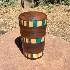 May include: A cylindrical wooden container with a lid, featuring alternating bands of dark brown walnut wood and decorative segments of light tan, orange, and teal blue wood. The container has a smooth, polished finish and a flat top surface.
