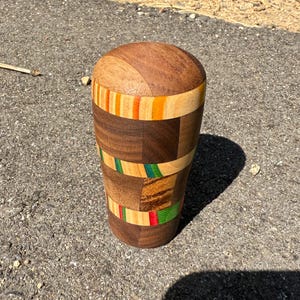 May include: A cylindrical wooden object featuring a rounded top and a segmented design. The body is composed of alternating blocks of dark brown wood, light tan wood, and thin horizontal bands of bright orange, green, and red colored wood.