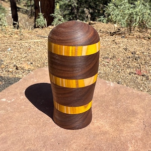 May include: A cylindrical wooden object with a rounded top, featuring alternating horizontal bands of dark brown walnut wood and bright yellow wood. The object has a smooth, polished finish and a slightly tapered shape toward the base.