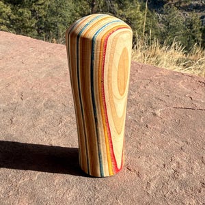 May include: A wooden shift knob with a cylindrical shape and a rounded top. The knob is made of layered wood in various colors, including red, blue, yellow, and natural wood tones. The shift knob is standing on a red rock surface.