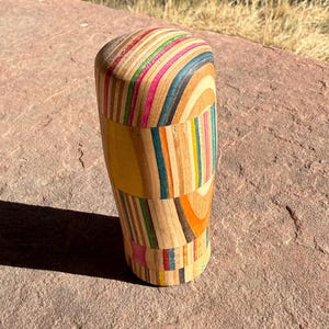 May include: A wooden, cylindrical object with a rounded top. The body is constructed from colorful, layered wood strips in shades of yellow, pink, blue, and orange. The object is sitting on a reddish-brown stone surface.