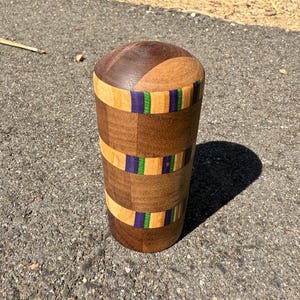 May include: A cylindrical wooden object featuring a segmented design with alternating bands of dark brown, light tan, purple, and bright green wood. The top is rounded and smooth, while the body consists of multiple small rectangular wood pieces.