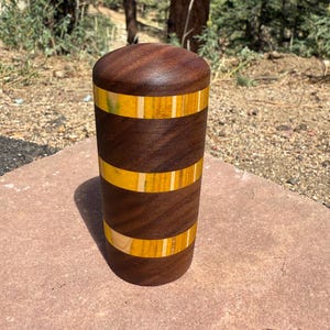 May include: A cylindrical wooden container with a rounded top, featuring alternating horizontal bands of dark brown walnut wood and bright yellow wood with thin light-colored accents, showcasing a smooth, polished finish and a segmented design.