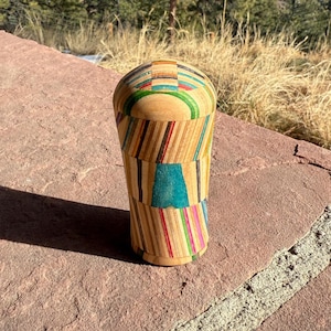 May include: A wooden, handcrafted cup with a unique design. The cup is made from small, colorful pieces of wood, creating a mosaic-like pattern. The colors include shades of blue, green, red, and yellow. The cup has a rounded top and a slightly tapered base.
