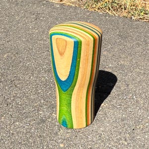 May include: A cylindrical gear shift knob crafted from laminated wood layers in natural tan, bright green, teal, and orange. The object features a smooth, rounded top and a tapered body with visible wood grain patterns throughout the vertical layers.