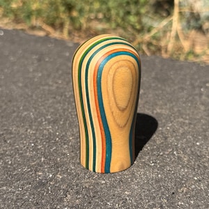 May include: A small, rounded, cylindrical object crafted from layered wood, featuring a natural light wood grain centre flanked by vertical stripes in shades of blue, orange, green, and yellow, creating a smooth, polished, and multi-coloured finish.
