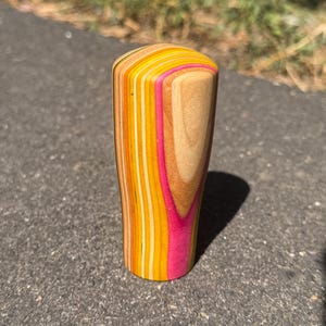 May include: A cylindrical gear shift knob crafted from layered wood, featuring a vertical pattern of natural light wood grain alternating with vibrant stripes of bright yellow, orange, and hot pink, with a smooth, rounded top surface.