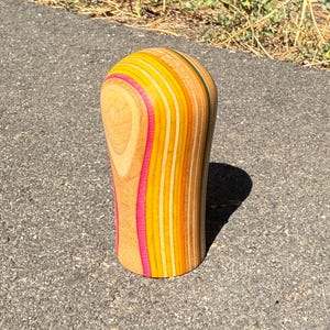May include: A rounded, upright wooden object crafted from multiple laminated layers of wood. The surface features a vertical pattern of natural wood grain alternating with vibrant stripes of hot pink, bright yellow, orange, and teal green.