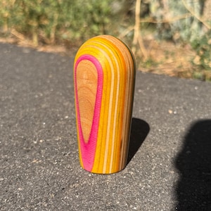 May include: A small, rounded, cylindrical object crafted from layered wood. The exterior features a vertical, bright neon pink stripe centered on a natural wood grain panel, surrounded by alternating bands of yellow and light tan wood layers.