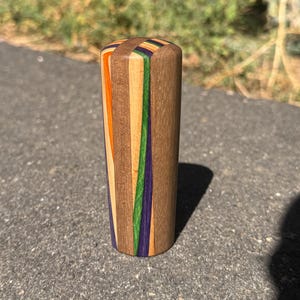 May include: A cylindrical wooden handle featuring a vertical, multi-coloured laminated design. The object is constructed from alternating strips of natural light brown wood, dark brown wood, bright orange, vibrant green, and deep purple wood.