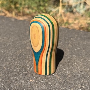 May include: A cylindrical wooden object with a rounded top, featuring vertical layers of natural light wood grain alternating with vibrant stripes of teal, orange, and green. The surface is smooth and polished, showcasing a multi-colored laminated pattern.