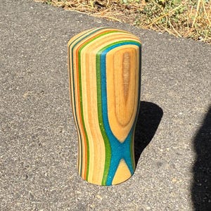 May include: A cylindrical gear shift knob crafted from laminated wood layers featuring vertical stripes in natural wood tones, bright blue, green, and orange. The top is rounded, and the body tapers slightly toward the base with a smooth finish.