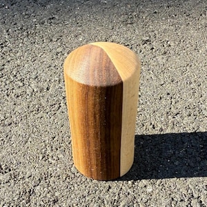 May include: A cylindrical wooden object with a rounded top. The object is made of two types of wood, with a dark brown section and a lighter tan section. The object is standing upright on a rough, gray surface.