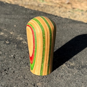 May include: A wooden shift knob with a rounded top and a cylindrical base. The knob features alternating layers of natural wood, green, and red, creating a striped pattern. The knob is designed for a vehicle's gear shift.